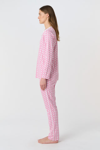 A woman stands barefoot against a plain background, wearing Roller Rabbit's Hathi Pajamas—a matching pink and white set with long sleeves and pants. She faces forward, smiling softly.