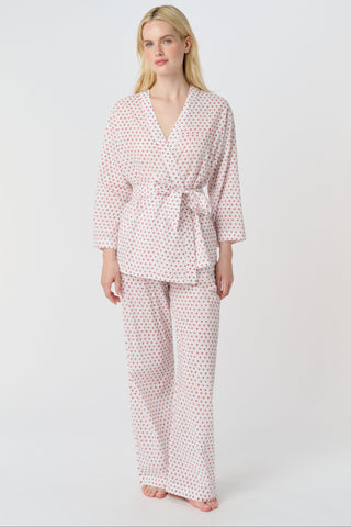 A person stands barefoot in the Roller Rabbit Hearts Christy Set—a light pink lounge set with small red hearts, featuring a belted wrap top and loose-fitting pants—against a plain white background.