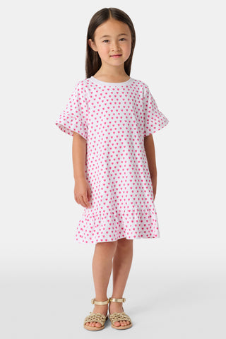 A young girl stands smiling in the Roller Rabbit Girls Hearts Esme Dress, a casual cotton T-shirt dress with pink heart patterns, short ruffled sleeves, and ruffled trim. She wears gold sandals and has long brown hair against a plain light background.