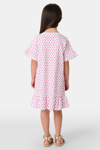 A young girl stands smiling in the Roller Rabbit Girls Hearts Esme Dress, a casual cotton T-shirt dress with pink heart patterns, short ruffled sleeves, and ruffled trim. She wears gold sandals and has long brown hair against a plain light background.