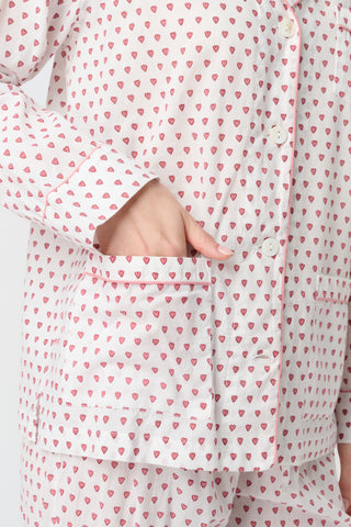A person with long blonde hair stands barefoot, wearing Roller Rabbit's Hearts Loungewear pajama set—a block-printed cotton ensemble with a light background and small red and blue dots, featuring a buttoned collared top with front pockets.