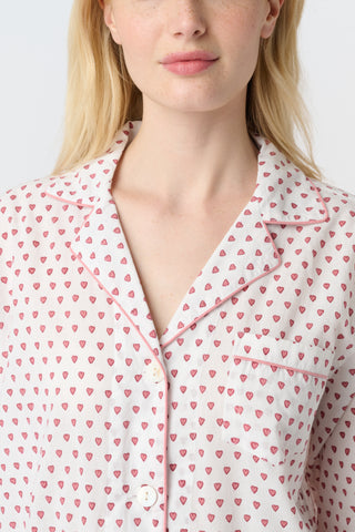 A person with long blonde hair stands barefoot, wearing Roller Rabbit's Hearts Loungewear pajama set—a block-printed cotton ensemble with a light background and small red and blue dots, featuring a buttoned collared top with front pockets.