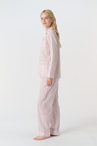 A person with long blonde hair stands barefoot, wearing Roller Rabbit's Hearts Loungewear pajama set—a block-printed cotton ensemble with a light background and small red and blue dots, featuring a buttoned collared top with front pockets.