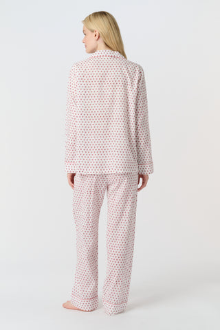 A person with long blonde hair stands barefoot, wearing Roller Rabbit's Hearts Loungewear pajama set—a block-printed cotton ensemble with a light background and small red and blue dots, featuring a buttoned collared top with front pockets.