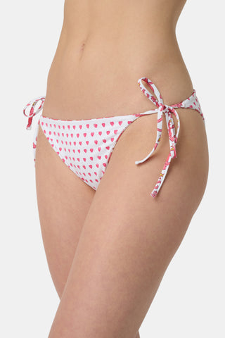 Someone wears the Roller Rabbit Monkey/Hearts Serena Bikini Bottom, a white reversible bikini bottom with pink and red animal and floral patterns, side ties, and UPF 50+ protection. Upper body and hips visible; plain white background.