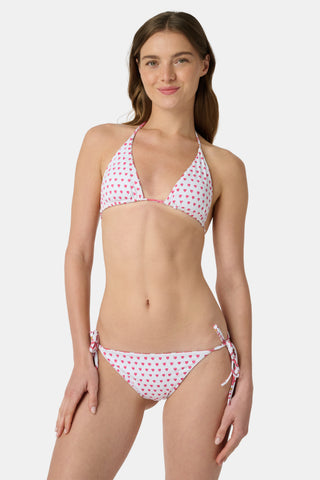 A woman in a straw hat and the Roller Rabbit Monkey/Hearts Serena Bikini Top smiles and leans forward at the beach, with ocean and sky behind her. The UPF 50+ fabric of this reversible bikini top offers sun protection.