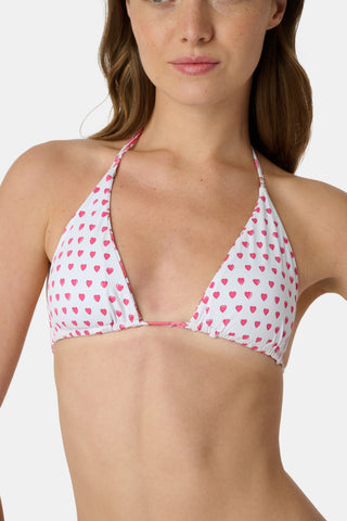 A woman in a straw hat and the Roller Rabbit Monkey/Hearts Serena Bikini Top smiles and leans forward at the beach, with ocean and sky behind her. The UPF 50+ fabric of this reversible bikini top offers sun protection.