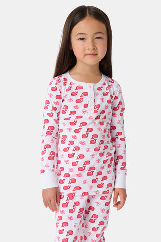 A young girl with curly hair stands barefoot, wearing Kids Moby Pajamas featuring a pink and red pattern. She faces forward with a relaxed expression against a plain white background.