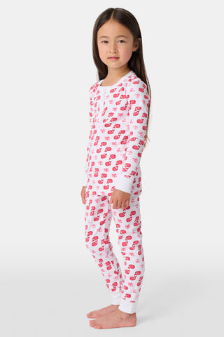 A young girl with curly hair stands barefoot, wearing Kids Moby Pajamas featuring a pink and red pattern. She faces forward with a relaxed expression against a plain white background.