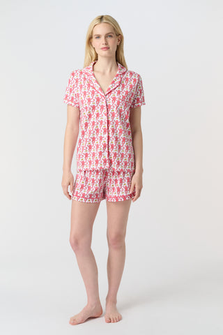 A woman with curly hair wears Monkey Polo Pajamas—a matching pima cotton short-sleeve set—featuring a pink and red crab print, standing against a matching background, smiling with her arms raised.