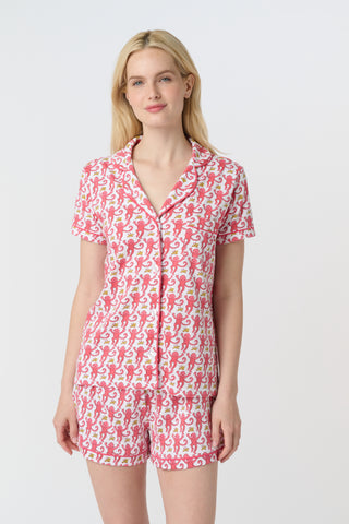 A woman with curly hair wears Monkey Polo Pajamas—a matching pima cotton short-sleeve set—featuring a pink and red crab print, standing against a matching background, smiling with her arms raised.
