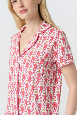 A woman with curly hair wears Monkey Polo Pajamas—a matching pima cotton short-sleeve set—featuring a pink and red crab print, standing against a matching background, smiling with her arms raised.