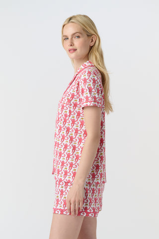 A woman with curly hair wears Monkey Polo Pajamas—a matching pima cotton short-sleeve set—featuring a pink and red crab print, standing against a matching background, smiling with her arms raised.