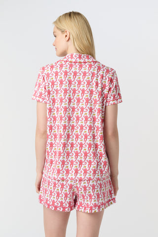 A woman with curly hair wears Monkey Polo Pajamas—a matching pima cotton short-sleeve set—featuring a pink and red crab print, standing against a matching background, smiling with her arms raised.
