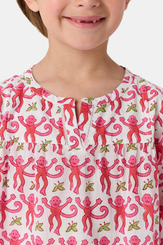 A young girl with brown hair smiles and stands against a white background wearing the Roller Rabbit Kids Monkey Serafina Tunic, a white cotton voile nightgown featuring a playful monkey print and ruffled hem.