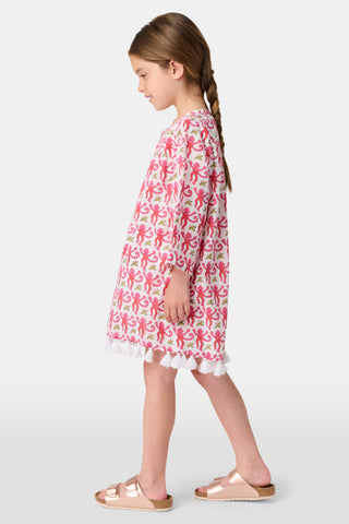 A young girl with brown hair smiles and stands against a white background wearing the Roller Rabbit Kids Monkey Serafina Tunic, a white cotton voile nightgown featuring a playful monkey print and ruffled hem.