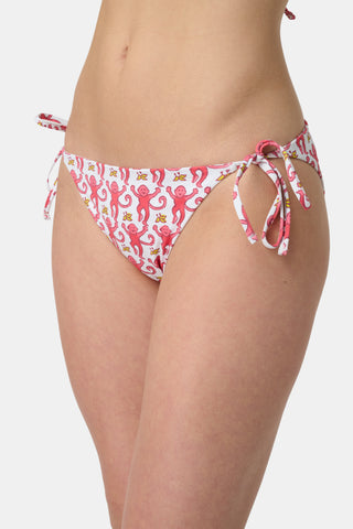 Someone wears the Roller Rabbit Monkey/Hearts Serena Bikini Bottom, a white reversible bikini bottom with pink and red animal and floral patterns, side ties, and UPF 50+ protection. Upper body and hips visible; plain white background.