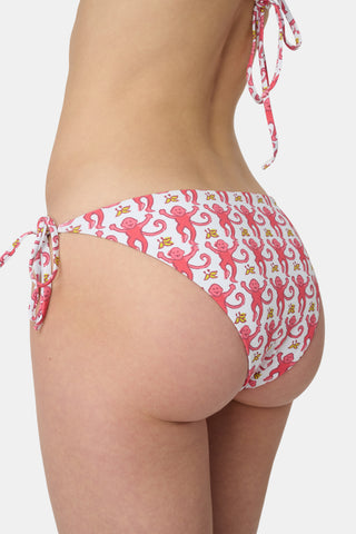 Someone wears the Roller Rabbit Monkey/Hearts Serena Bikini Bottom, a white reversible bikini bottom with pink and red animal and floral patterns, side ties, and UPF 50+ protection. Upper body and hips visible; plain white background.
