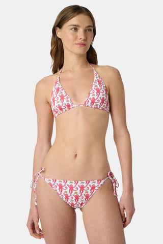 A woman in a straw hat and the Roller Rabbit Monkey/Hearts Serena Bikini Top smiles and leans forward at the beach, with ocean and sky behind her. The UPF 50+ fabric of this reversible bikini top offers sun protection.