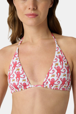 A woman in a straw hat and the Roller Rabbit Monkey/Hearts Serena Bikini Top smiles and leans forward at the beach, with ocean and sky behind her. The UPF 50+ fabric of this reversible bikini top offers sun protection.