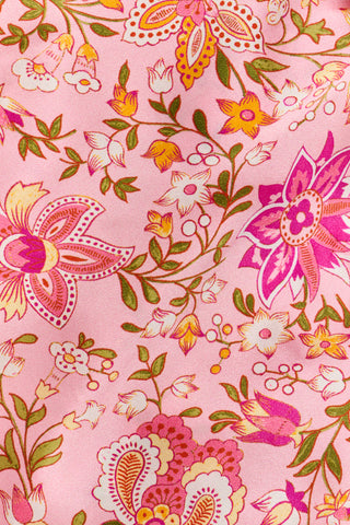A woman stands barefoot against a plain white background wearing the Priya Floral Alvie Set by Roller Rabbit—a matching pink pajama set featuring a floral pattern, short-sleeve button-up top, and loose pants crafted from washable silk loungewear.