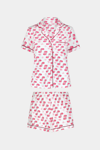 A woman wears Roller Rabbit's Moby Polo Pajamas—a women's short sleeve set with a white base and pink-red cartoon fruit prints, button-up top, and shorts. She stands against a plain light background, smiling gently.