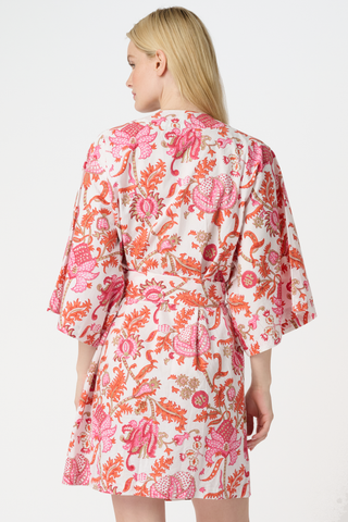 A woman with long blonde hair wears the Roller Rabbit Amanda Kimono, a belted, knee-length cotton kimono with a vibrant pink and orange floral pattern on white. She stands against a plain light gray backdrop, smiling softly.