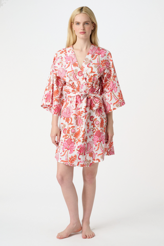 A woman with long blonde hair wears the Roller Rabbit Amanda Kimono, a belted, knee-length cotton kimono with a vibrant pink and orange floral pattern on white. She stands against a plain light gray backdrop, smiling softly.