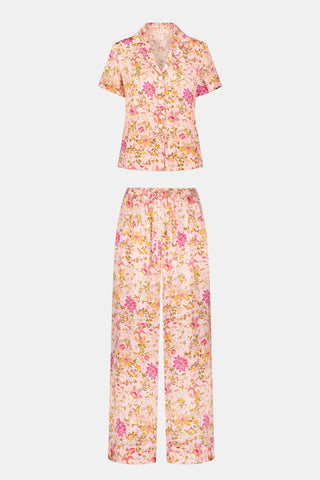 A woman stands barefoot against a plain white background wearing the Priya Floral Alvie Set by Roller Rabbit—a matching pink pajama set featuring a floral pattern, short-sleeve button-up top, and loose pants crafted from washable silk loungewear.