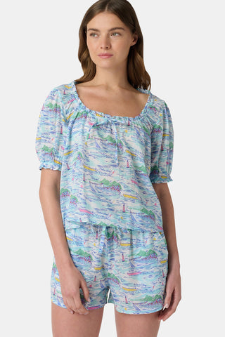 A woman stands facing forward wearing the Roller Rabbit Regatta Preston Set, a light blue and pastel-patterned women's two-piece with short sleeves and matching shorts, paired with white sandals against a plain white background.