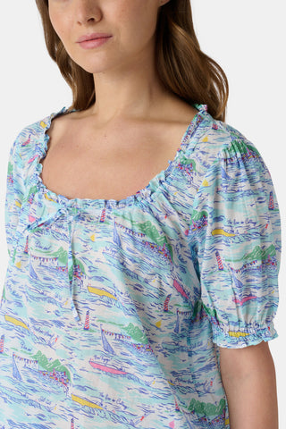 A woman stands facing forward wearing the Roller Rabbit Regatta Preston Set, a light blue and pastel-patterned women's two-piece with short sleeves and matching shorts, paired with white sandals against a plain white background.