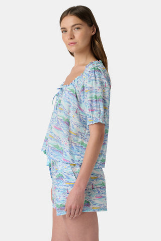 A woman stands facing forward wearing the Roller Rabbit Regatta Preston Set, a light blue and pastel-patterned women's two-piece with short sleeves and matching shorts, paired with white sandals against a plain white background.
