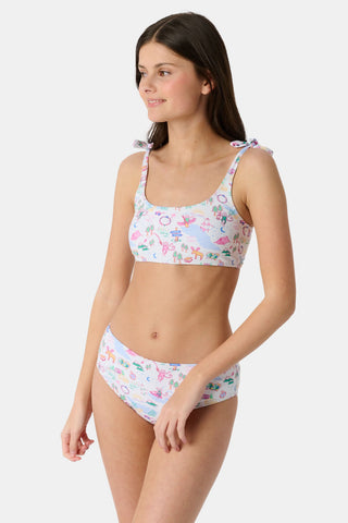 A girl wears the Roller Rabbit Kids Camp RR Cora Bikini, featuring a colorful pattern, adjustable tie straps, and high-waisted bottoms. She smiles against a white background, highlighting the comfort and UV protection of this swimwear.