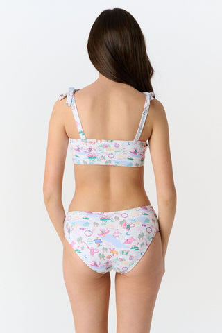 A girl wears the Roller Rabbit Kids Camp RR Cora Bikini, featuring a colorful pattern, adjustable tie straps, and high-waisted bottoms. She smiles against a white background, highlighting the comfort and UV protection of this swimwear.