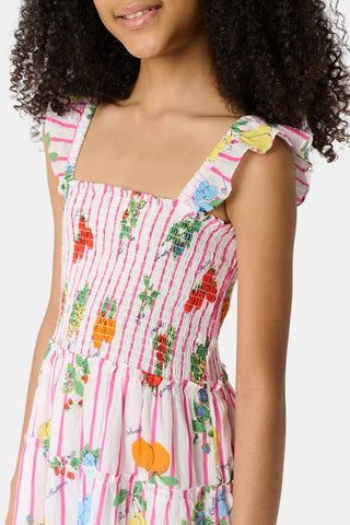 A young girl with straight brown hair wears the Roller Rabbit Kids Fruits D'Ete Ceri Dress, a pink and white smocked sundress with ruffled sleeves, and white sandals, standing and smiling against a light gray background.