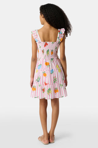 A young girl with straight brown hair wears the Roller Rabbit Kids Fruits D'Ete Ceri Dress, a pink and white smocked sundress with ruffled sleeves, and white sandals, standing and smiling against a light gray background.