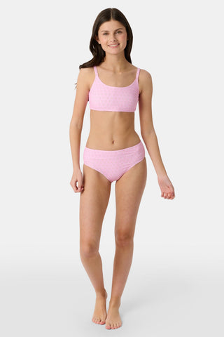 A teen girl stands smiling, wearing the Roller Rabbit Kids Hearts Lola Bikini—a light pink two-piece swimsuit with small heart patterns and full coverage bottoms—against a plain light gray background.