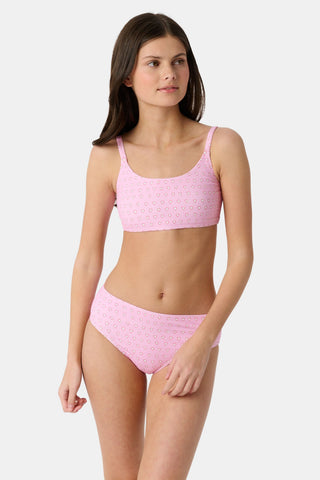 A teen girl stands smiling, wearing the Roller Rabbit Kids Hearts Lola Bikini—a light pink two-piece swimsuit with small heart patterns and full coverage bottoms—against a plain light gray background.