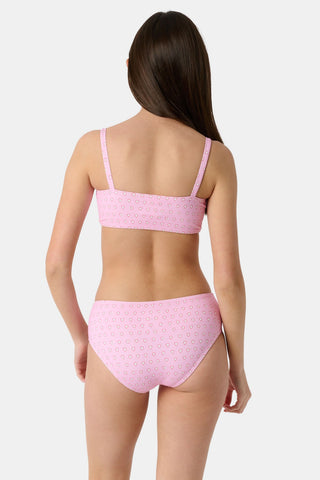 A teen girl stands smiling, wearing the Roller Rabbit Kids Hearts Lola Bikini—a light pink two-piece swimsuit with small heart patterns and full coverage bottoms—against a plain light gray background.