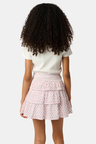 A young girl with curly hair, seen from behind, wears a white short-sleeve top and the Kids Hearts Sami Skirt—a pink ruffle mini skirt with a hearts print—standing against a plain light background.