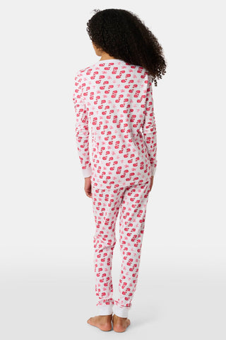 A child with curly hair stands barefoot, facing away, in Kids Moby Pajamas—a white set with pink and red Santa prints. Made from soft Pima cotton, the pajamas stand out against the plain white background.