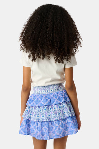 A young girl with curly hair wears a plain white t-shirt and the blue Roller Rabbit Kids Monkey/Jemina Sami Skirt with a smocked waist, standing with her hands behind her back against a plain, light background.