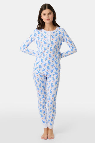 A young child with curly hair stands barefoot in white Kids Romy Rabbit Pajamas by Roller Rabbit, featuring a blue bunny print, looking at the camera and touching their mouth with one finger.