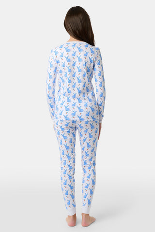 A young child with curly hair stands barefoot in white Kids Romy Rabbit Pajamas by Roller Rabbit, featuring a blue bunny print, looking at the camera and touching their mouth with one finger.
