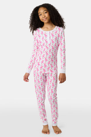 A young girl smiles barefoot in Roller Rabbit’s Kids Romy the Rabbit Pajamas, a soft Pima cotton long-sleeve set with a playful rabbit print; her brown hair frames her face against a simple, light background.