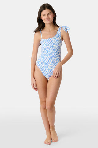A young girl with long brown hair wears the Roller Rabbit Kids Seaside Knots Jacey One Piece, featuring a blue geometric pattern and a bow on one shoulder, smiling with her hands on her hips against a plain background.