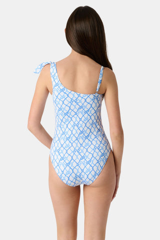 A young girl with long brown hair wears the Roller Rabbit Kids Seaside Knots Jacey One Piece, featuring a blue geometric pattern and a bow on one shoulder, smiling with her hands on her hips against a plain background.