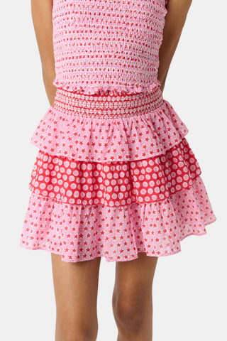 A child wears the Roller Rabbit Kids Starry Dot Sami Skirt in pink and red with star and polka dot patterns, paired with a pink smocked top, standing against a plain light background.