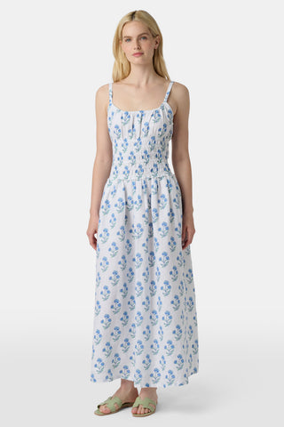 A woman wears the Roller Rabbit Rosa Floral Veronica Dress, a white sundress with blue prints, a smocked bodice, and spaghetti straps, styled with light green sandals as she stands facing forward with her arms relaxed by her sides.