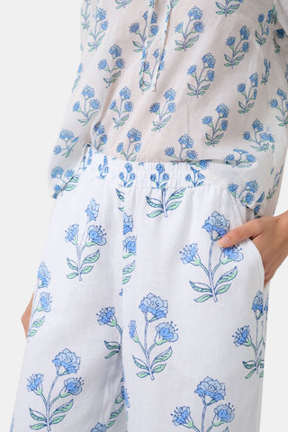A person wears the Roller Rabbit Rosa Floral Kaylee Pant—high-waisted white linen pants with blue floral print and pockets—paired with an off-white top and green sandals, standing against a plain light background.